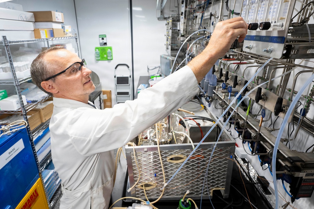 A male laboratory technician or scientist in a white lab coat and safety glasses is adjusting equipment on a complex panel of valves, tubing, and instrumentation. The setting appears to be a professional laboratory or pilot plant environment with multiple fluid handling systems. The image captures active hands-on technical work with sophisticated process equipment.