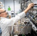 A male laboratory technician or scientist in a white lab coat and safety glasses is adjusting equipment on a complex panel of valves, tubing, and instrumentation. The setting appears to be a professional laboratory or pilot plant environment with multiple fluid handling systems. The image captures active hands-on technical work with sophisticated process equipment.