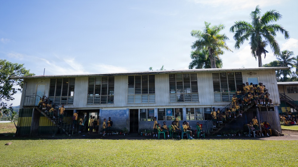 A two-story school building in what appears to be a Pacific Island or tropical setting, surrounded by palm trees and lush green grass. Large numbers of students wearing matching yellow and green uniforms are gathered outside the building, sitting on stairs, standing on the upper balcony, and congregating at ground level. The building has a weathered, utilitarian appearance with louvered windows and a corrugated metal roof.