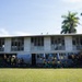 A two-story school building in what appears to be a Pacific Island or tropical setting, surrounded by palm trees and lush green grass. Large numbers of students wearing matching yellow and green uniforms are gathered outside the building, sitting on stairs, standing on the upper balcony, and congregating at ground level. The building has a weathered, utilitarian appearance with louvered windows and a corrugated metal roof.