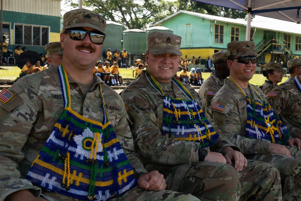 Three U.S. Army soldiers are seated and smiling, wearing camouflage uniforms and traditional woven decorative garments in blue, green, yellow, and white colors, likely gifted during a cultural exchange or community engagement event. In the background, a large group of children in yellow school uniforms are gathered near green-painted buildings. The setting appears to be an outdoor ceremony or event in a tropical location.