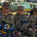 Three U.S. Army soldiers are seated and smiling, wearing camouflage uniforms and traditional woven decorative garments in blue, green, yellow, and white colors, likely gifted during a cultural exchange or community engagement event. In the background, a large group of children in yellow school uniforms are gathered near green-painted buildings. The setting appears to be an outdoor ceremony or event in a tropical location.