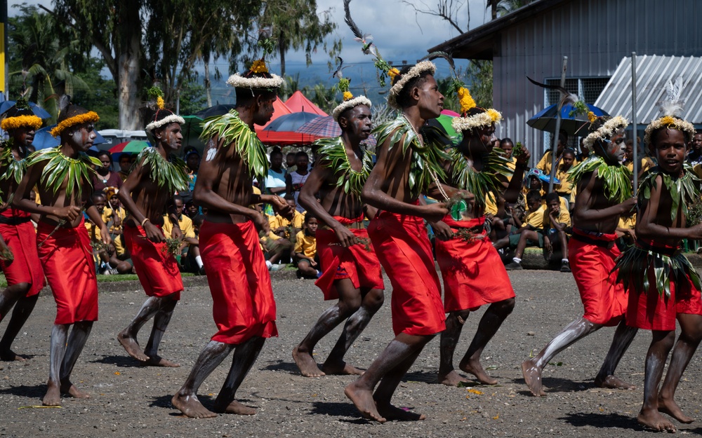 A group of traditional dancers perform in coordinated formation wearing red wrap skirts, leaf shoulder decorations, and floral headdresses. The performance takes place outdoors on a gravel surface before a large seated audience wearing yellow shirts. The setting appears to be a cultural festival or ceremonial event in a Pacific Island or Melanesian context.