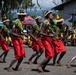 A group of traditional dancers perform in coordinated formation wearing red wrap skirts, leaf shoulder decorations, and floral headdresses. The performance takes place outdoors on a gravel surface before a large seated audience wearing yellow shirts. The setting appears to be a cultural festival or ceremonial event in a Pacific Island or Melanesian context.