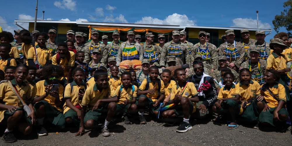 A group photo featuring military personnel in camouflage uniforms posing with a large group of children wearing yellow and green school uniforms. The setting appears to be outdoors in front of a yellow and green building decorated with orange and yellow bunting, suggesting a ceremonial or community event. The military personnel are wearing colorful traditional garlands/leis, indicating a cultural exchange or goodwill visit.