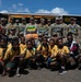 A group photo featuring military personnel in camouflage uniforms posing with a large group of children wearing yellow and green school uniforms. The setting appears to be outdoors in front of a yellow and green building decorated with orange and yellow bunting, suggesting a ceremonial or community event. The military personnel are wearing colorful traditional garlands/leis, indicating a cultural exchange or goodwill visit.