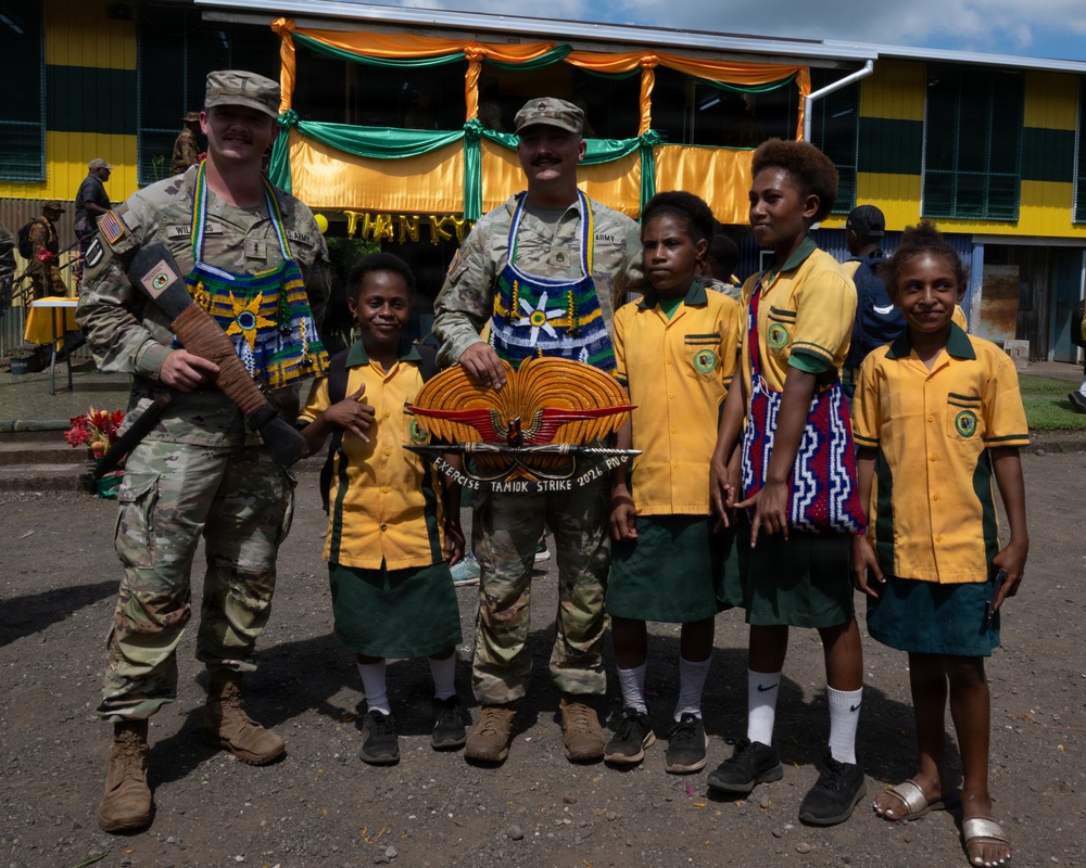 Two U.S. Army soldiers wearing traditional Papua New Guinean beaded necklaces/bilas pose with four local schoolchildren in front of a decorated building. The center soldier holds a decorative plaque reading 'Exercise Tamiok Strike 2026 PNG.' This appears to be a community engagement event during a joint military exercise in Papua New Guinea.