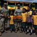 Two U.S. Army soldiers wearing traditional Papua New Guinean beaded necklaces/bilas pose with four local schoolchildren in front of a decorated building. The center soldier holds a decorative plaque reading 'Exercise Tamiok Strike 2026 PNG.' This appears to be a community engagement event during a joint military exercise in Papua New Guinea.