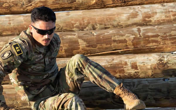 A soldier in full camouflage uniform is navigating a log obstacle course, appearing to climb or traverse horizontally stacked wooden logs. The individual wears tactical gear including gloves, sunglasses, and tan combat boots, with a number '42' bib visible on their left arm. The image captures an athletic training or competition event, likely a military fitness challenge or obstacle course race.