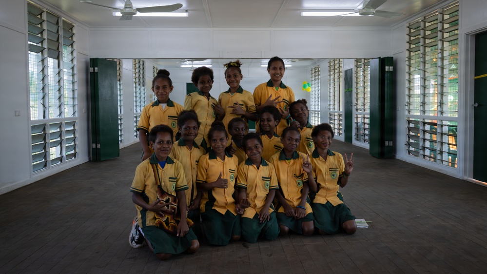 A group of approximately 12 school girls are posing together for a group photo inside what appears to be a school building or hall. They are all wearing matching yellow and green school uniforms. The students are arranged in rows, with some sitting on the floor and others standing behind them.