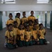 A group of approximately 12 school girls are posing together for a group photo inside what appears to be a school building or hall. They are all wearing matching yellow and green school uniforms. The students are arranged in rows, with some sitting on the floor and others standing behind them.