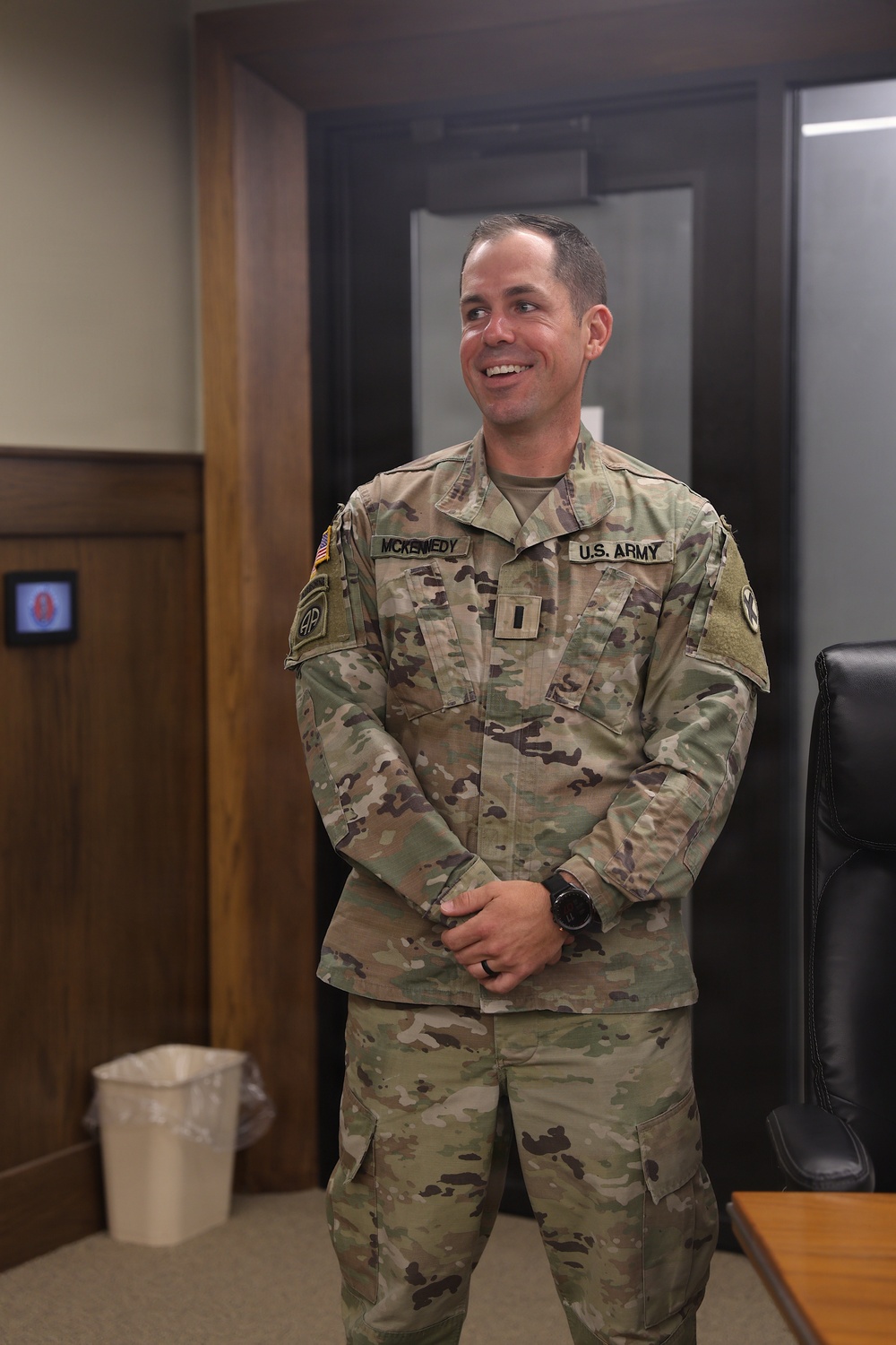A U.S. Army soldier wearing Operational Camouflage Pattern (OCP) uniform is standing in what appears to be an office or administrative building interior. The soldier is smiling and looking to his left, with hands clasped in front. His name tape reads 'MCKENNEDY' and rank insignia indicates he is a Captain (O-3).