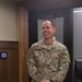 A U.S. Army soldier wearing Operational Camouflage Pattern (OCP) uniform is standing in what appears to be an office or administrative building interior. The soldier is smiling and looking to his left, with hands clasped in front. His name tape reads 'MCKENNEDY' and rank insignia indicates he is a Captain (O-3).