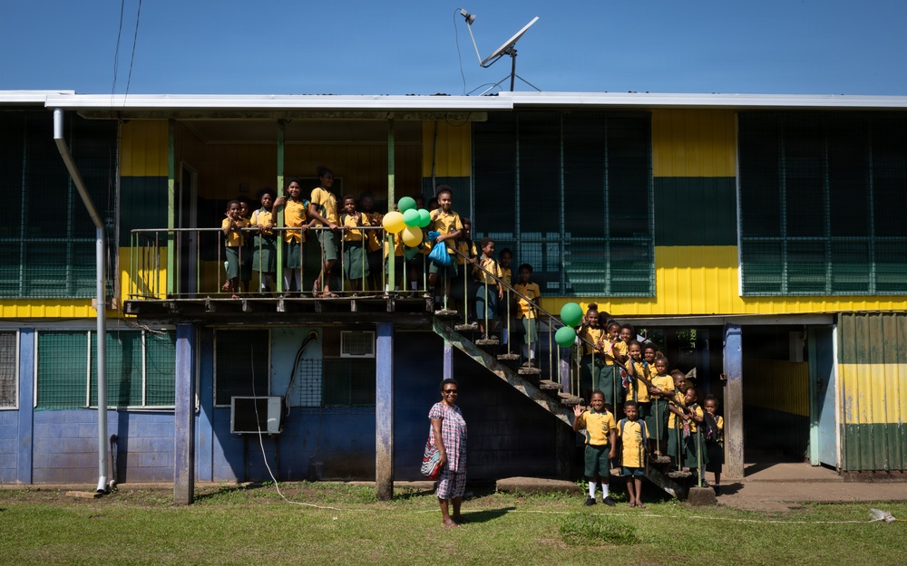 A group of school children in matching yellow and green uniforms are gathered on the balcony and staircase of a two-story school building painted in yellow, green, and blue. An adult woman in a patterned dress stands on the ground in front of the building. Some children are holding green and yellow balloons, suggesting a celebratory occasion.