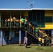 A group of school children in matching yellow and green uniforms are gathered on the balcony and staircase of a two-story school building painted in yellow, green, and blue. An adult woman in a patterned dress stands on the ground in front of the building. Some children are holding green and yellow balloons, suggesting a celebratory occasion.