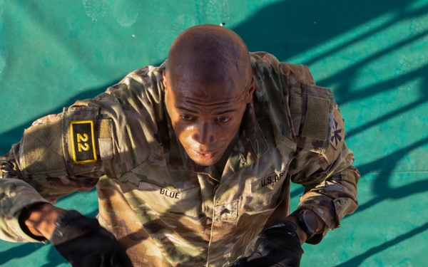 A U.S. Army soldier named 'Blue' is navigating a rope obstacle course or cargo net, wearing OCP (Operational Camouflage Pattern) multicam uniform with black tactical gloves. The soldier's uniform displays a unit patch with the number '22' on the left shoulder and a rank insignia on the chest. The image appears to capture a physical training or obstacle course event in an outdoor setting.