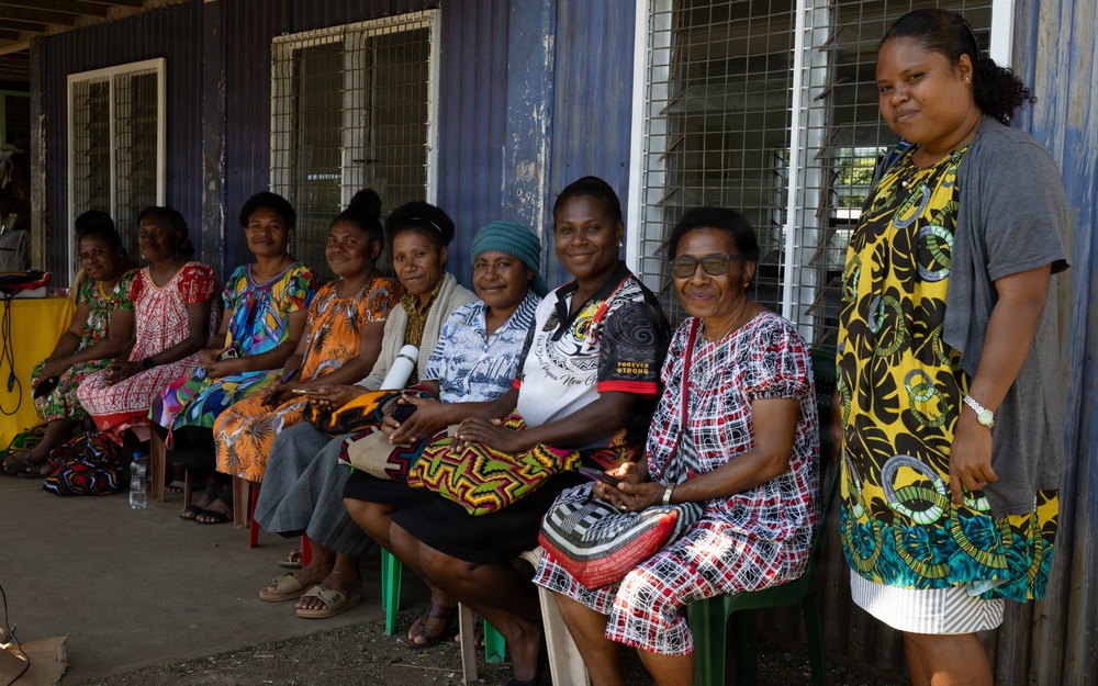A group of approximately nine women are gathered along the exterior wall of a blue corrugated metal building, most seated on plastic chairs in a row while one stands at the right. The women are dressed in colorful, patterned clothing typical of Pacific Island communities, suggesting this may be Papua New Guinea or a nearby region. The setting appears to be a community or public building, and the group seems to be assembled for a meeting or event.