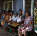 A group of approximately nine women are gathered along the exterior wall of a blue corrugated metal building, most seated on plastic chairs in a row while one stands at the right. The women are dressed in colorful, patterned clothing typical of Pacific Island communities, suggesting this may be Papua New Guinea or a nearby region. The setting appears to be a community or public building, and the group seems to be assembled for a meeting or event.