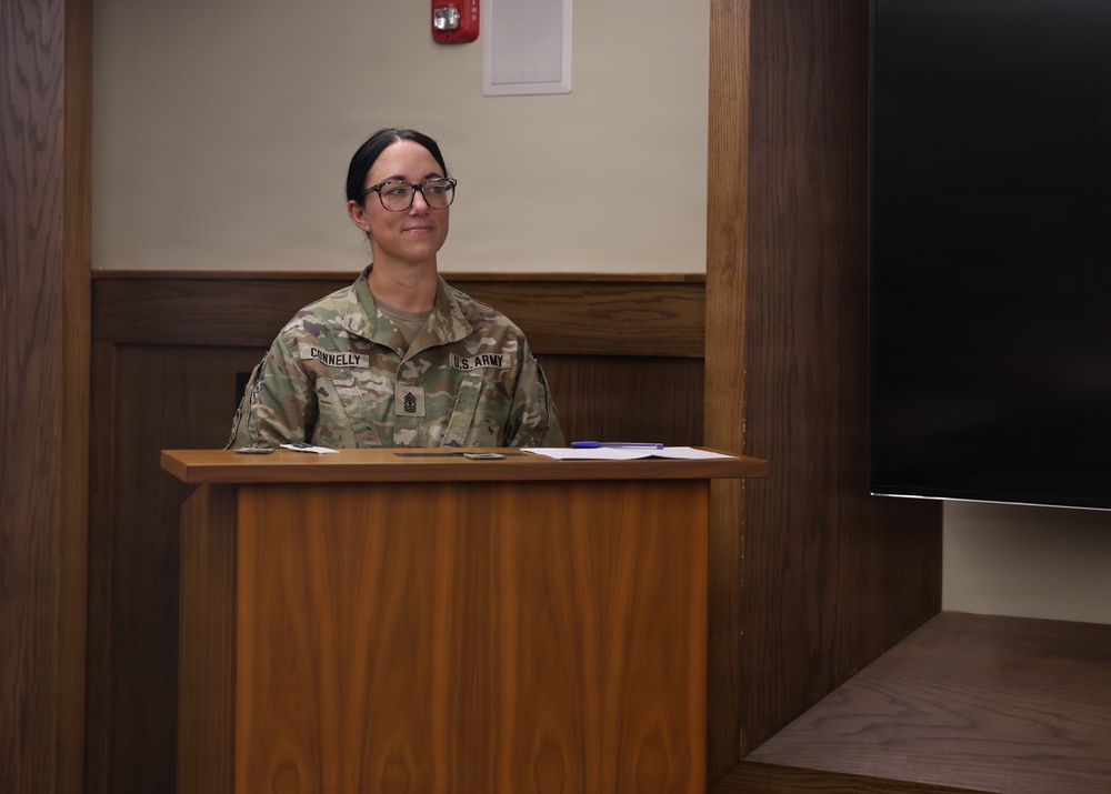 A U.S. Army soldier named Connelly, wearing OCP (Operational Camouflage Pattern) uniform, stands at a wooden podium in what appears to be a formal briefing room or conference facility. The soldier's rank insignia on the chest indicates a senior non-commissioned officer grade. Papers and small items are visible on the podium surface.