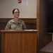 A U.S. Army soldier named Connelly, wearing OCP (Operational Camouflage Pattern) uniform, stands at a wooden podium in what appears to be a formal briefing room or conference facility. The soldier's rank insignia on the chest indicates a senior non-commissioned officer grade. Papers and small items are visible on the podium surface.