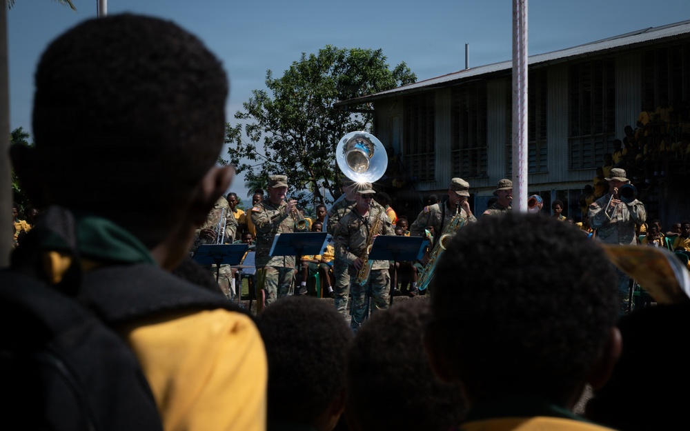 Military personnel in camouflage uniforms are performing as a brass band for a crowd of children, likely at a school or community event. The audience, predominantly children wearing yellow uniforms, watches the performance attentively. The setting appears to be an outdoor courtyard of a building in what appears to be a Pacific Island or African nation.