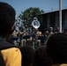 Military personnel in camouflage uniforms are performing as a brass band for a crowd of children, likely at a school or community event. The audience, predominantly children wearing yellow uniforms, watches the performance attentively. The setting appears to be an outdoor courtyard of a building in what appears to be a Pacific Island or African nation.