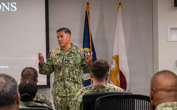A U.S. Navy officer in NWU Type III (woodland camouflage) uniform is delivering a briefing or presentation to a group of seated Navy personnel in a conference room setting. A projected slide behind him displays the partial word 'CTATIONS' (likely 'EXPECTATIONS') along with text referencing 'Navy Medicine' and rank standards. Two flags are visible in the background, consistent with a U.S. military command environment.