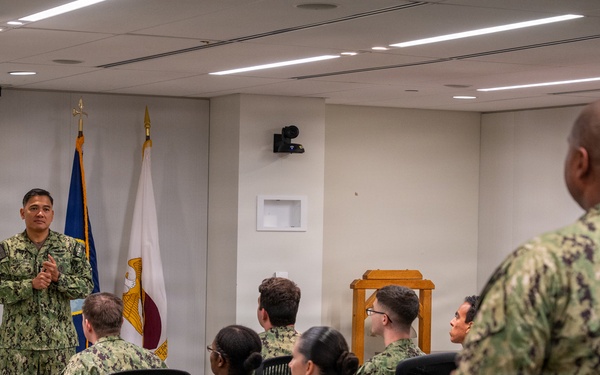 A military briefing or presentation is taking place in a modern conference room, with one uniformed service member standing at the front addressing a seated group. All individuals are wearing U.S. Navy Working Uniform (NWU Type III) camouflage pattern. Two flags are visible at the front of the room alongside a projection screen.