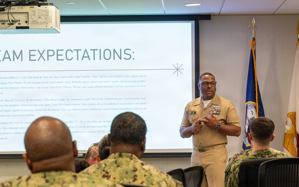 A senior U.S. Navy officer in Service Khaki uniform is delivering a briefing or leadership presentation to a group of sailors seated in a modern conference room. The projected slide behind him displays 'TEAM EXPECTATIONS' with detailed text outlining leadership responsibilities by enlisted rank. The audience is wearing Navy Working Uniform (NWU) camouflage pattern.