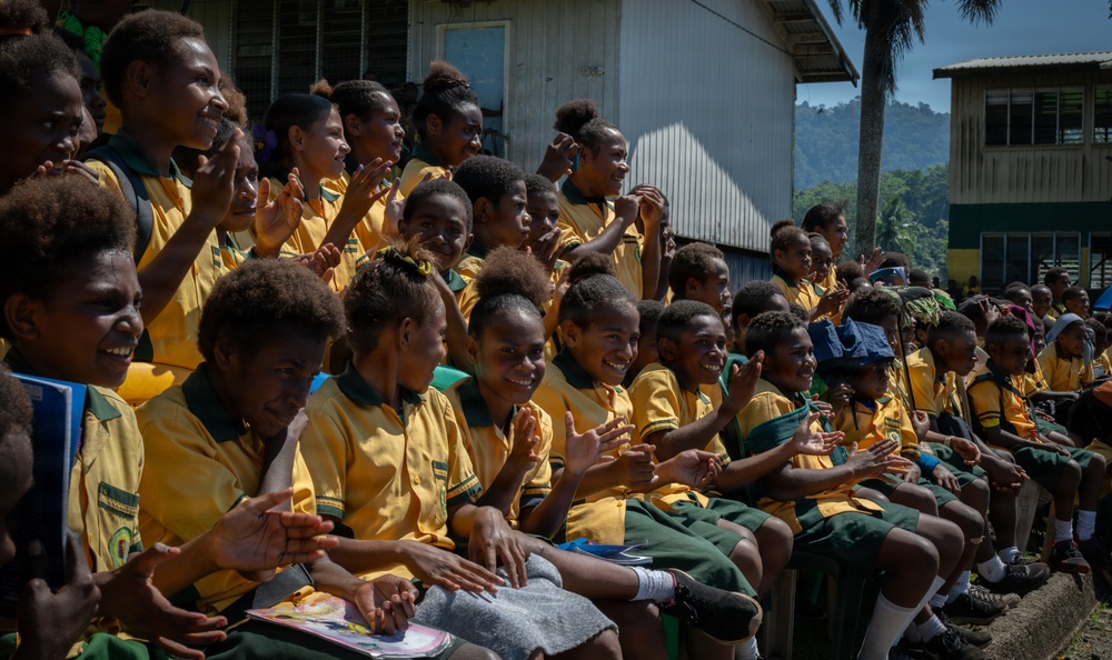A large group of school children wearing matching yellow and green uniforms are seated together outdoors, likely at a school event or assembly. The children appear joyful and engaged, with many smiling, clapping, and waving. The setting includes school buildings and tropical vegetation in the background, suggesting a Pacific Island or Southeast Asian location.