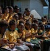 A large group of school children wearing matching yellow and green uniforms are seated together outdoors, likely at a school event or assembly. The children appear joyful and engaged, with many smiling, clapping, and waving. The setting includes school buildings and tropical vegetation in the background, suggesting a Pacific Island or Southeast Asian location.
