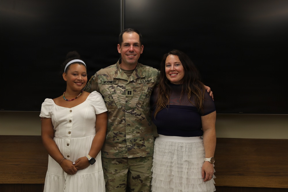 A U.S. Army soldier in OCP (Operational Camouflage Pattern) uniform poses with two civilians, likely family members, in what appears to be an indoor military or institutional setting. The soldier's name tape reads 'MCKENNEDY' and displays a Captain's rank insignia. The group appears to be posing for a formal or celebratory photo.