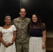 A U.S. Army soldier in OCP (Operational Camouflage Pattern) uniform poses with two civilians, likely family members, in what appears to be an indoor military or institutional setting. The soldier's name tape reads 'MCKENNEDY' and displays a Captain's rank insignia. The group appears to be posing for a formal or celebratory photo.