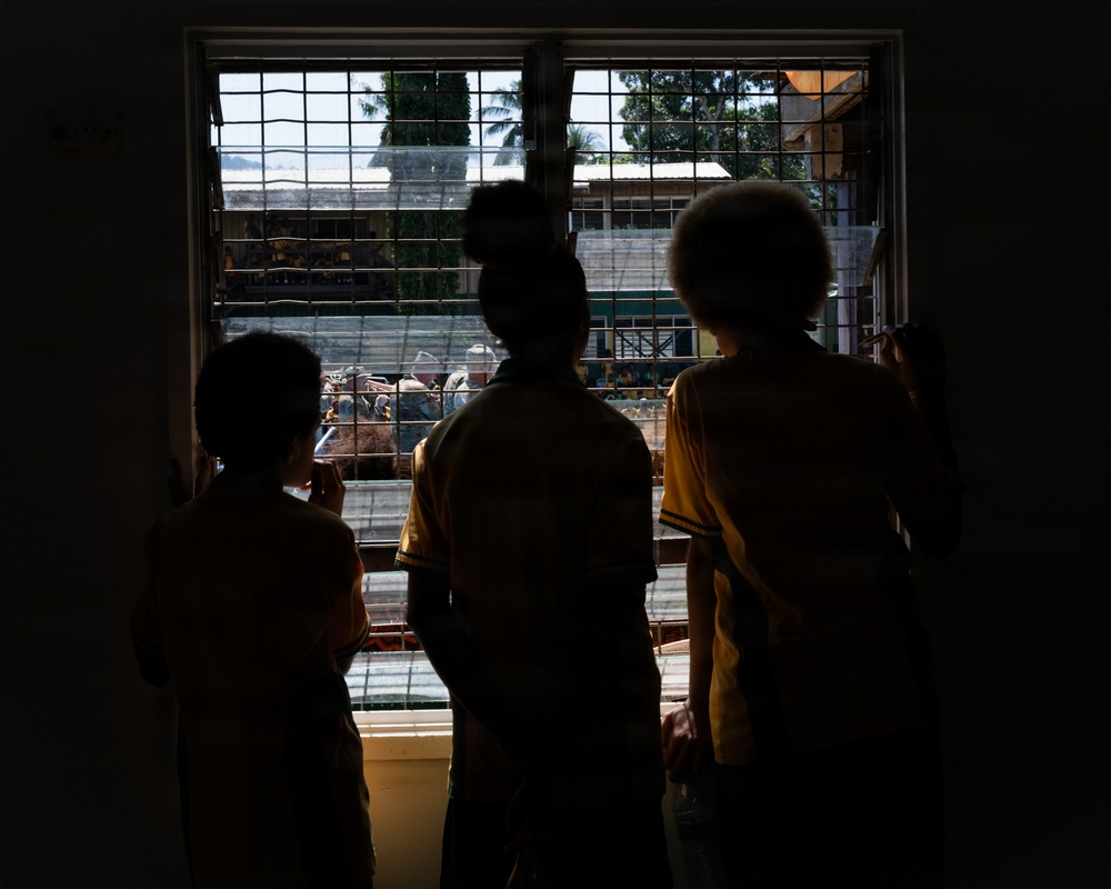 Three children are silhouetted against a barred window, looking out at an outdoor scene below. The backlit composition creates strong contrast between the dark interior and the bright exterior. The setting appears to be a school or institutional building based on the visible architecture and what appear to be school uniforms.