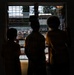 Three children are silhouetted against a barred window, looking out at an outdoor scene below. The backlit composition creates strong contrast between the dark interior and the bright exterior. The setting appears to be a school or institutional building based on the visible architecture and what appear to be school uniforms.