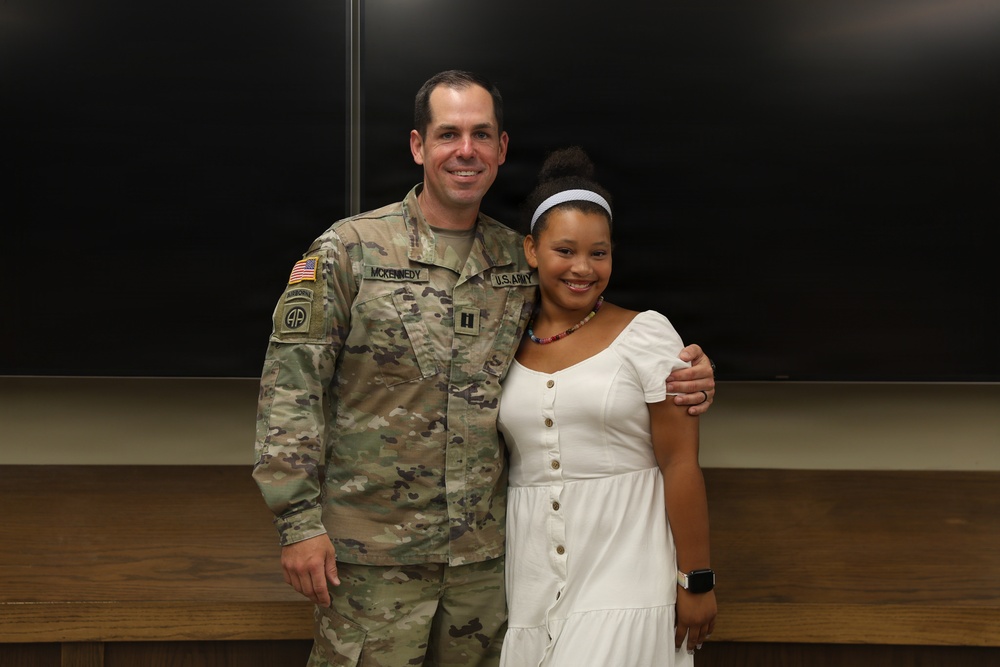 A U.S. Army soldier wearing OCP (Operational Camouflage Pattern) uniform with the name tape 'McKENNEY' and a Captain rank insignia poses with a young woman in a white dress. The soldier displays an 82nd Airborne Division patch on his left shoulder and a U.S. flag patch. They are standing in what appears to be a classroom or briefing room with chalkboards in the background.