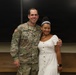 A U.S. Army soldier wearing OCP (Operational Camouflage Pattern) uniform with the name tape 'McKENNEY' and a Captain rank insignia poses with a young woman in a white dress. The soldier displays an 82nd Airborne Division patch on his left shoulder and a U.S. flag patch. They are standing in what appears to be a classroom or briefing room with chalkboards in the background.