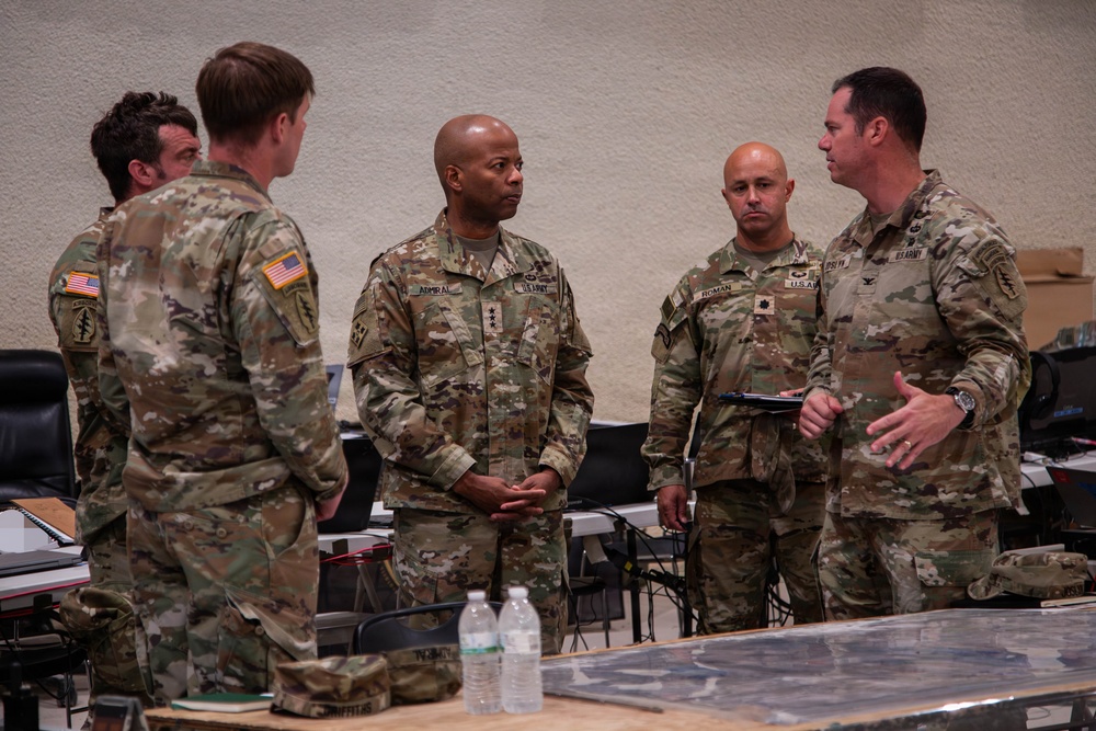 A group of five U.S. Army personnel in OCP (Operational Camouflage Pattern) uniforms are engaged in a briefing or discussion inside what appears to be a military operations center or command post. The central figure, identifiable by name tape 'ADMIRAL' and three-star rank insignia, appears to be a Lieutenant General receiving a briefing. Maps, laptops, and tactical equipment are visible on tables in the background.
