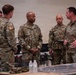 A group of five U.S. Army personnel in OCP (Operational Camouflage Pattern) uniforms are engaged in a briefing or discussion inside what appears to be a military operations center or command post. The central figure, identifiable by name tape 'ADMIRAL' and three-star rank insignia, appears to be a Lieutenant General receiving a briefing. Maps, laptops, and tactical equipment are visible on tables in the background.