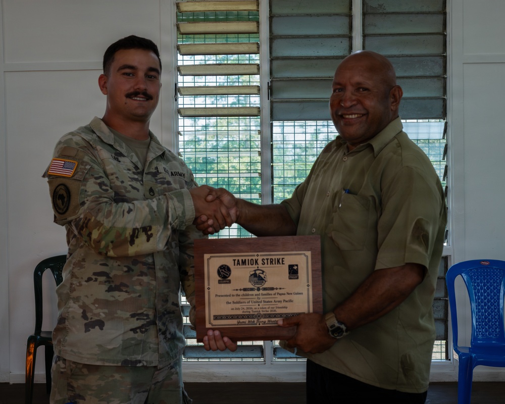A U.S. Army soldier in multicam uniform is shaking hands with a civilian man while both hold a wooden plaque. The plaque reads 'TAMIOK STRIKE' and references a presentation to the children and families of Papua New Guinea by the Soldiers of United States Army Pacific. The ceremony appears to be a formal recognition or goodwill exchange event.