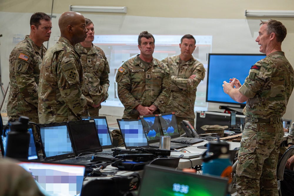 A group of U.S. Army soldiers in MultiCam/OCP camouflage uniforms are gathered in what appears to be a tactical operations center (TOC) or command post. One soldier on the right is briefing or presenting to the group, gesturing with his hands. Multiple laptops and computer monitors are arranged on tables in the foreground, suggesting a technology-intensive command environment.