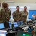 A group of U.S. Army soldiers in MultiCam/OCP camouflage uniforms are gathered in what appears to be a tactical operations center (TOC) or command post. One soldier on the right is briefing or presenting to the group, gesturing with his hands. Multiple laptops and computer monitors are arranged on tables in the foreground, suggesting a technology-intensive command environment.