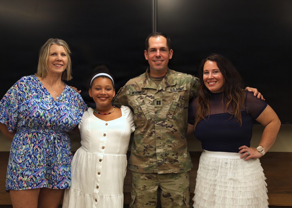 Four people pose together for a group photo, including a U.S. Army soldier in OCP (Operational Camouflage Pattern) uniform with the name tape 'McKENNEDY' and captain rank insignia visible. The group consists of three females and one male military member, appearing to be at an indoor ceremony or formal occasion. The atmosphere is celebratory and familial in nature.