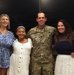 Four people pose together for a group photo, including a U.S. Army soldier in OCP (Operational Camouflage Pattern) uniform with the name tape 'McKENNEDY' and captain rank insignia visible. The group consists of three females and one male military member, appearing to be at an indoor ceremony or formal occasion. The atmosphere is celebratory and familial in nature.