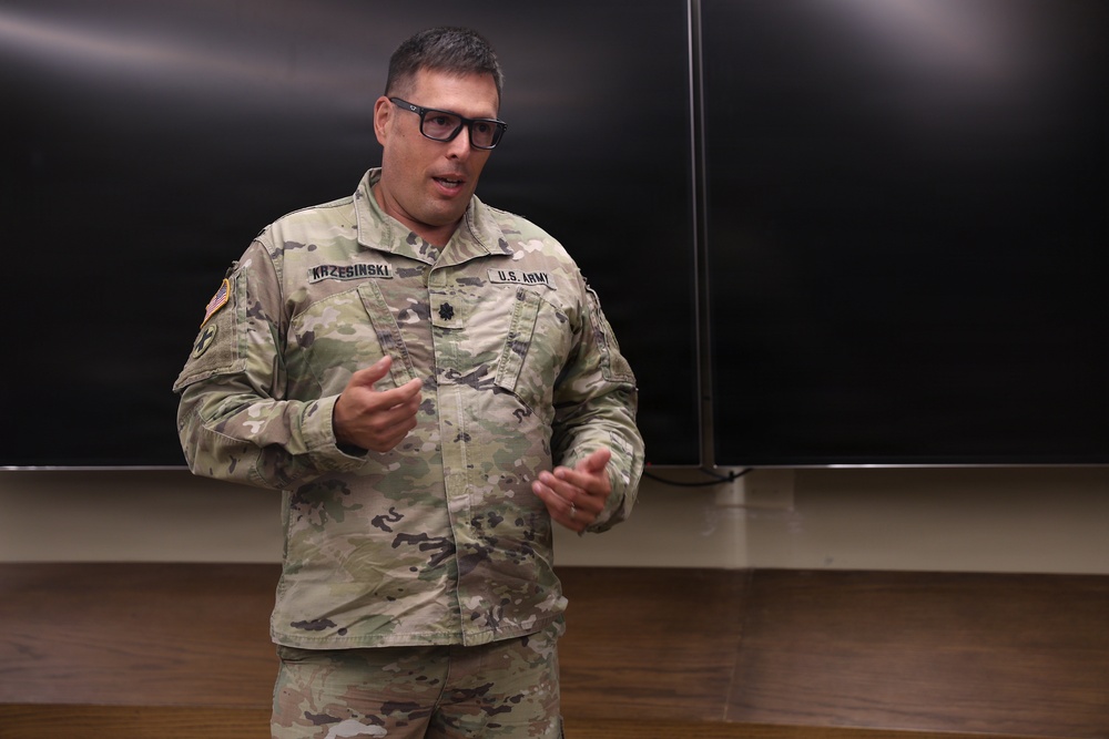 A U.S. Army officer wearing OCP (Operational Camouflage Pattern) uniform is speaking or presenting in what appears to be a classroom or briefing room setting. The individual's name tape reads 'KRZESINSKI' and wears a Lieutenant Colonel oak leaf cluster insignia on the chest. He is gesturing with both hands, suggesting active communication or instruction.