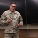 A U.S. Army officer wearing OCP (Operational Camouflage Pattern) uniform is speaking or presenting in what appears to be a classroom or briefing room setting. The individual's name tape reads 'KRZESINSKI' and wears a Lieutenant Colonel oak leaf cluster insignia on the chest. He is gesturing with both hands, suggesting active communication or instruction.