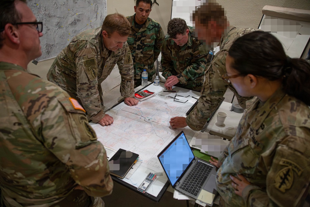 A group of military personnel in various camouflage uniforms are gathered around a table conducting a map briefing or planning session. Several faces have been intentionally pixelated/redacted for privacy. The group includes both U.S. Army soldiers and personnel in a different camouflage pattern, suggesting a joint or multinational planning exercise.
