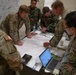 A group of military personnel in various camouflage uniforms are gathered around a table conducting a map briefing or planning session. Several faces have been intentionally pixelated/redacted for privacy. The group includes both U.S. Army soldiers and personnel in a different camouflage pattern, suggesting a joint or multinational planning exercise.