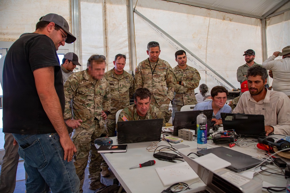A group of U.S. Army personnel in MultiCam/OCP camouflage uniforms and civilian contractors are gathered around a table with multiple laptops inside a large white tent or temporary structure. They appear to be engaged in a technical briefing or collaborative work session. The setting suggests a forward operating base or field exercise environment.
