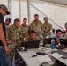 A group of U.S. Army personnel in MultiCam/OCP camouflage uniforms and civilian contractors are gathered around a table with multiple laptops inside a large white tent or temporary structure. They appear to be engaged in a technical briefing or collaborative work session. The setting suggests a forward operating base or field exercise environment.